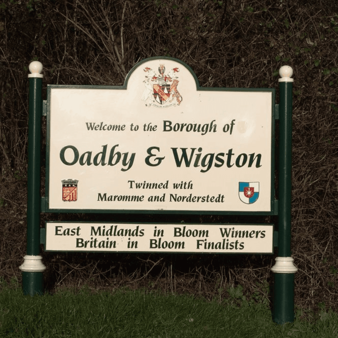 Sign reading "Welcome to the Borough of Oadby & Wigston, Twinned with Maromme and Norderstedt, Award Winners" in a grassy area. - Home Instead