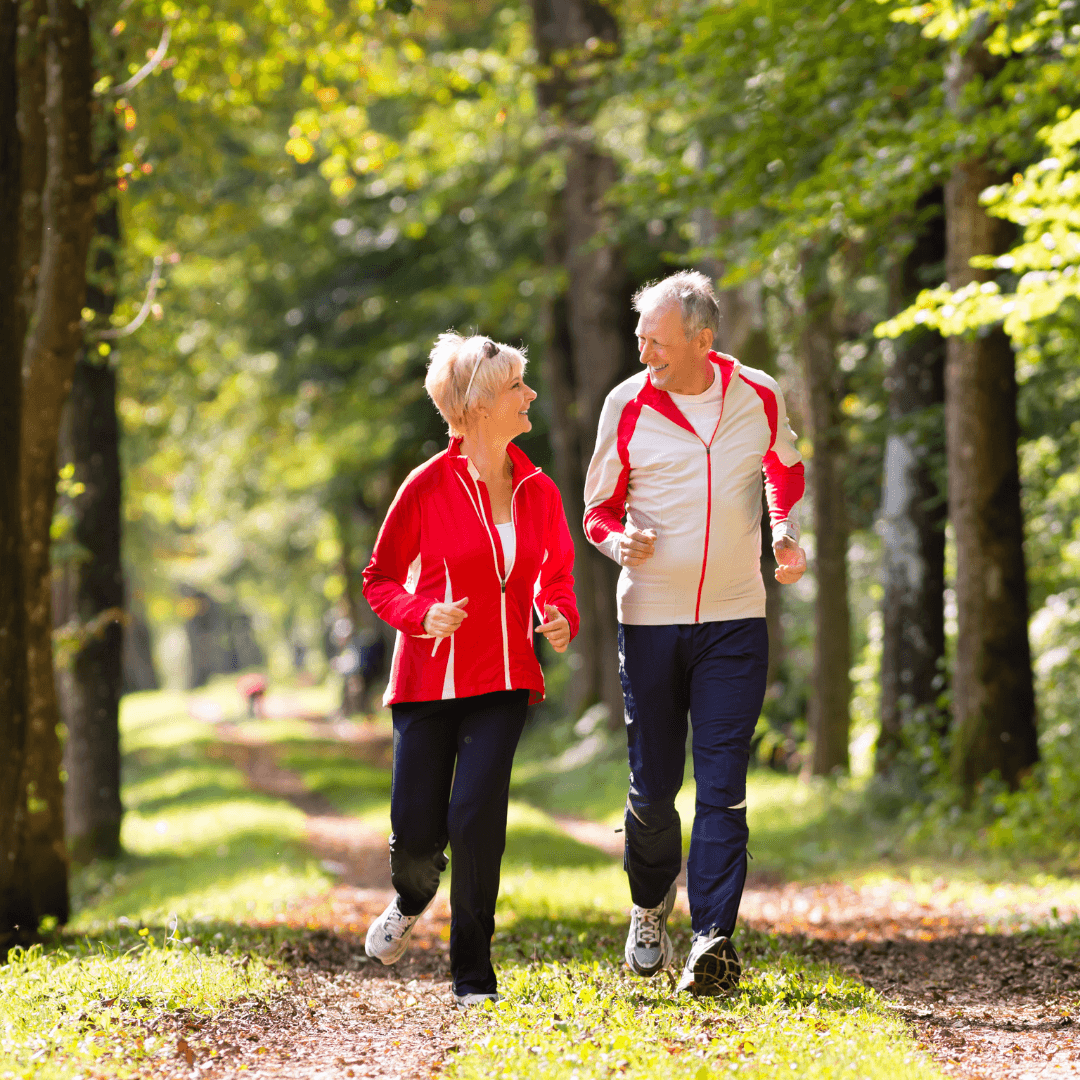An older couple in matching athletic wear jogs together on a wooded path, smiling and enjoying the outdoors. - Home Instead
