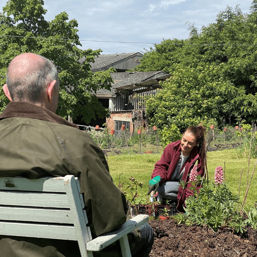A person in a jacket sits on a bench watching another person gardening in a lush, green area with trees and buildings. - Home Instead