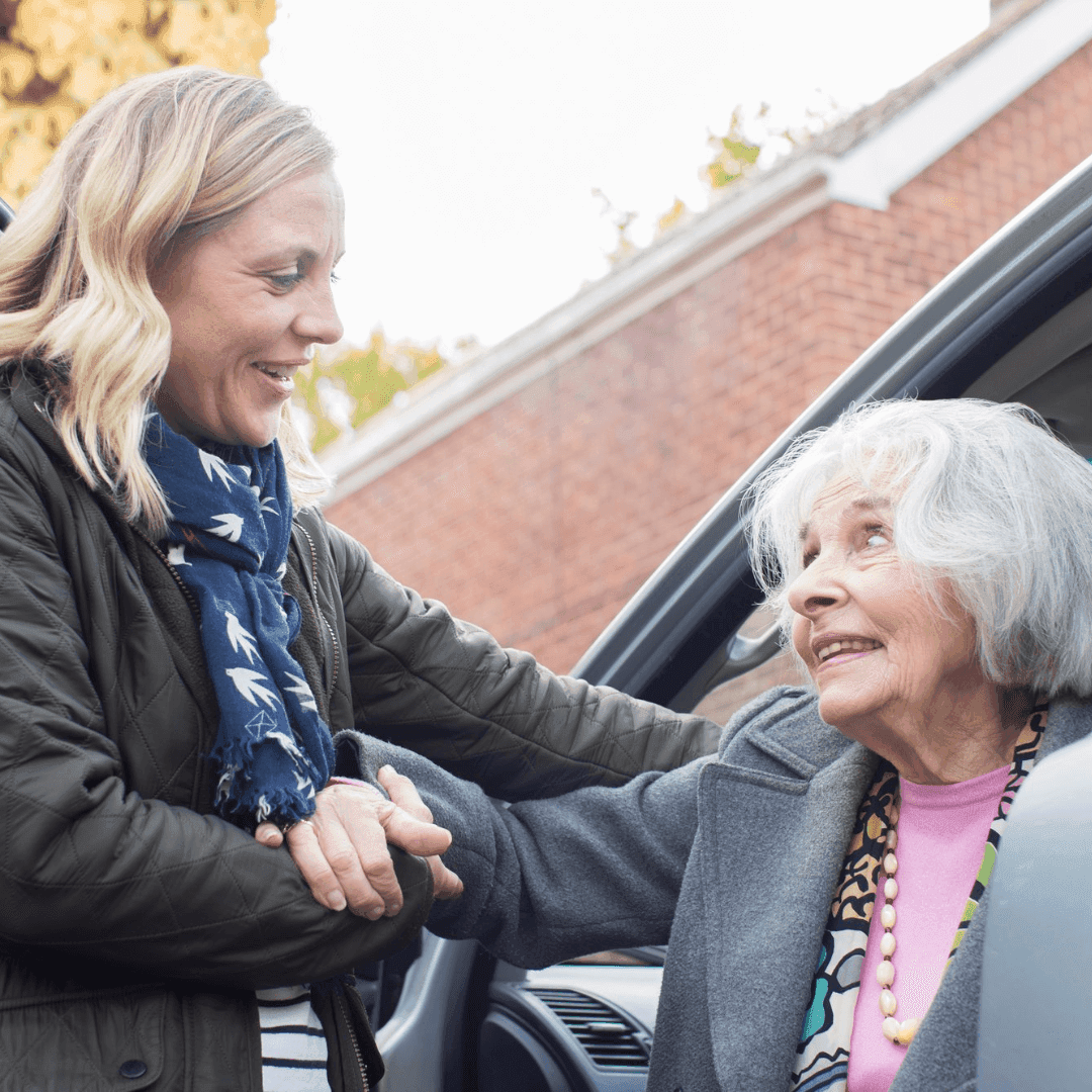 A woman helps an elderly woman out of a car, holding her hand and smiling warmly outdoors. - Home Instead