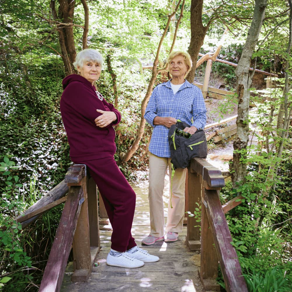 Two elderly women stand and smile on a wooden footbridge in a lush, green forest. - Home Instead