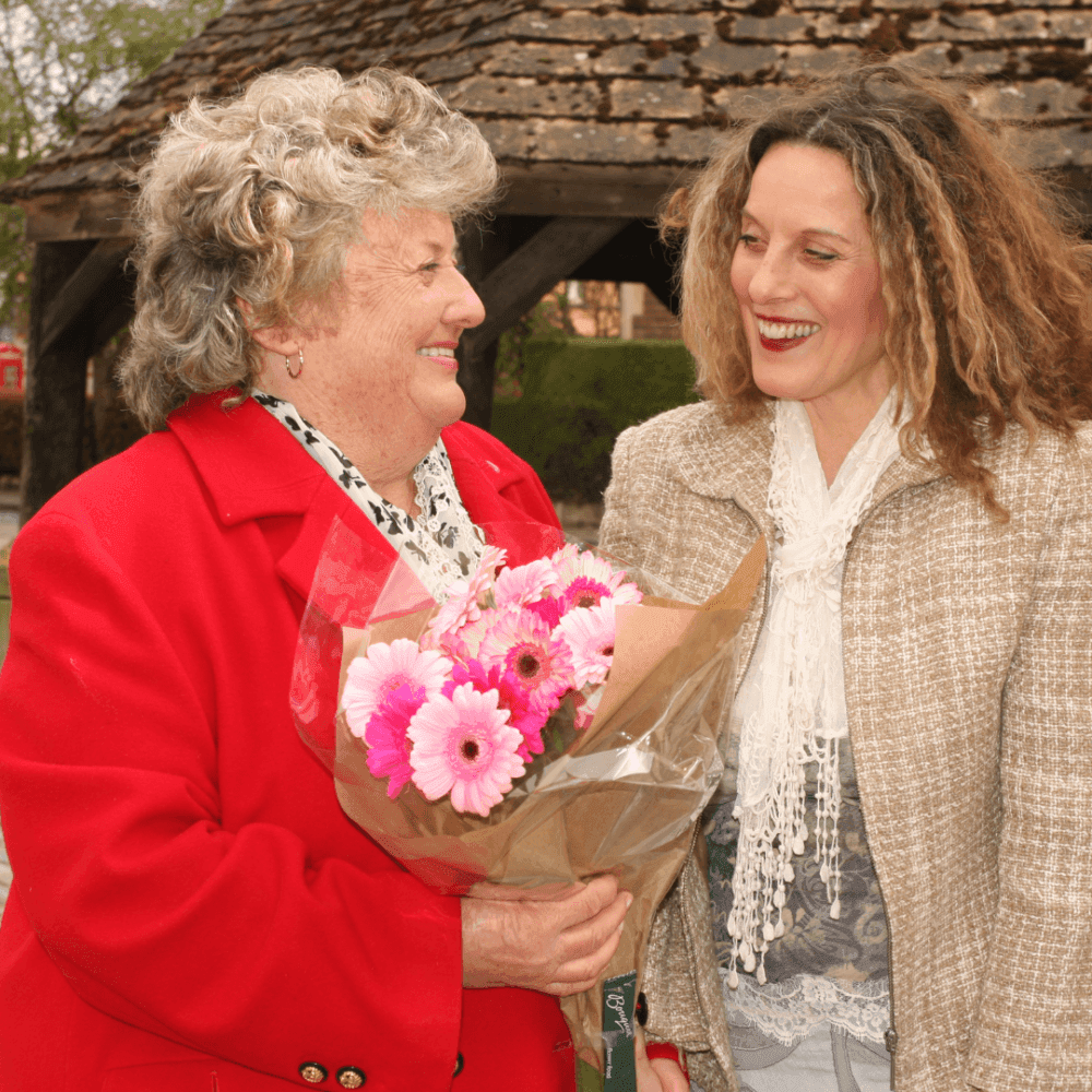 Two women, one holding a bouquet of pink flowers, smiling at each other outdoors. - Home Instead