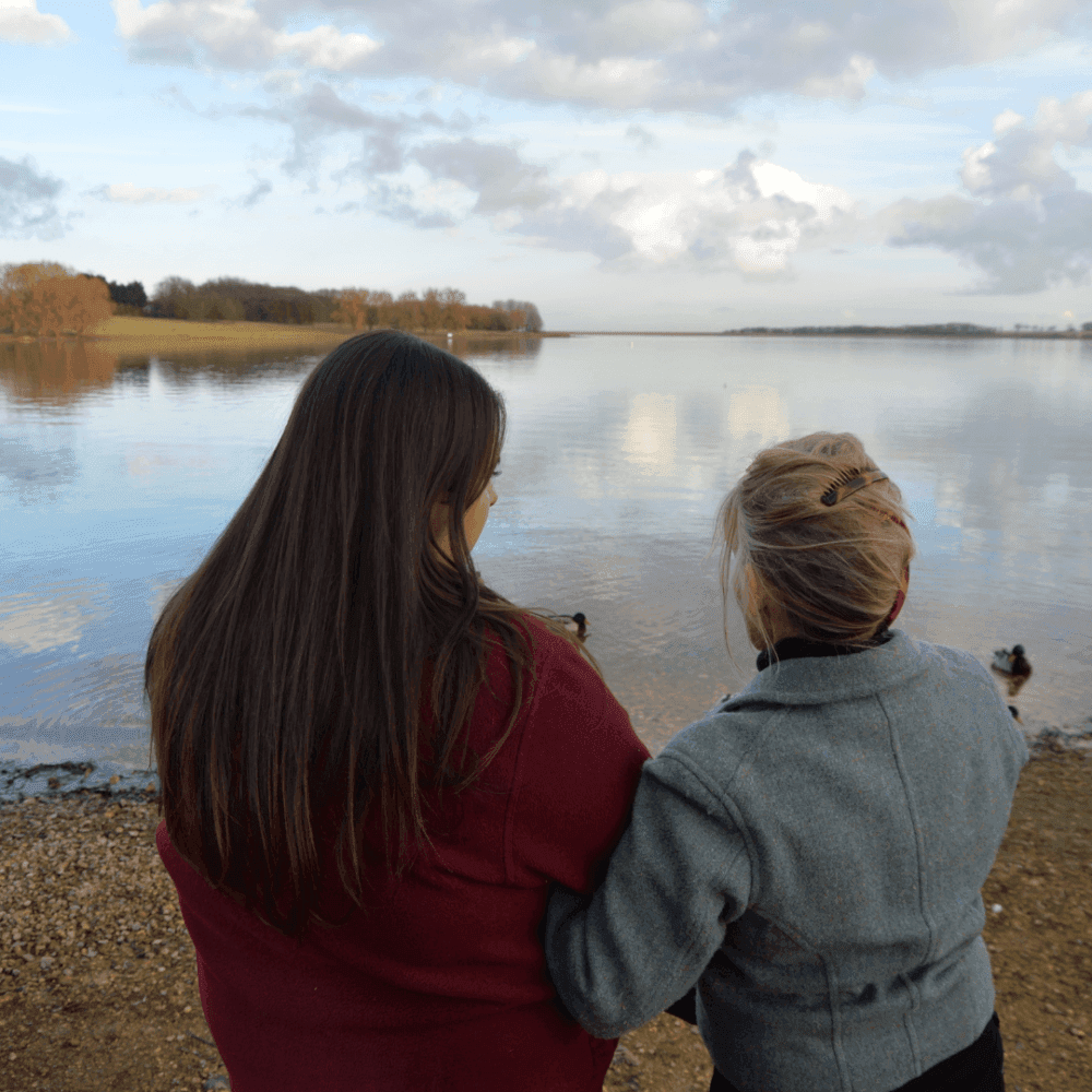Two women with an arm around each other, taking in a calm lake view on a partly cloudy day. - Home Instead