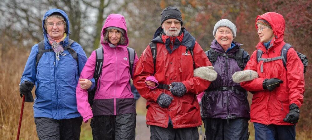 Five adults in colorful rain gear walk outdoors on a rainy day, smiling and enjoying the hike. - Home Instead