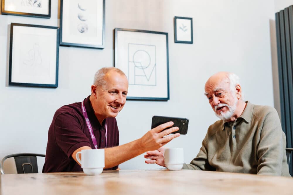 Two elderly men smiling and looking at a smartphone while sitting at a table with coffee mugs in a room with framed art. - Home Instead