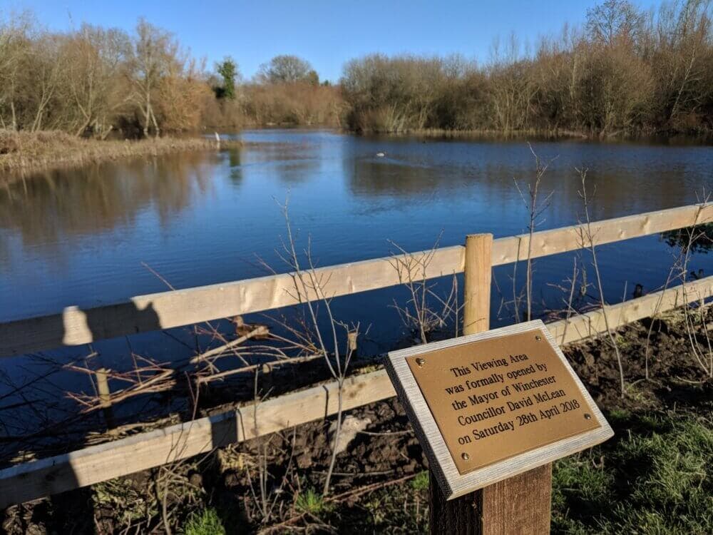 A wooden fence by a serene pond with a plaque about the Mayor of Winchester opening the viewing area. - Home Instead