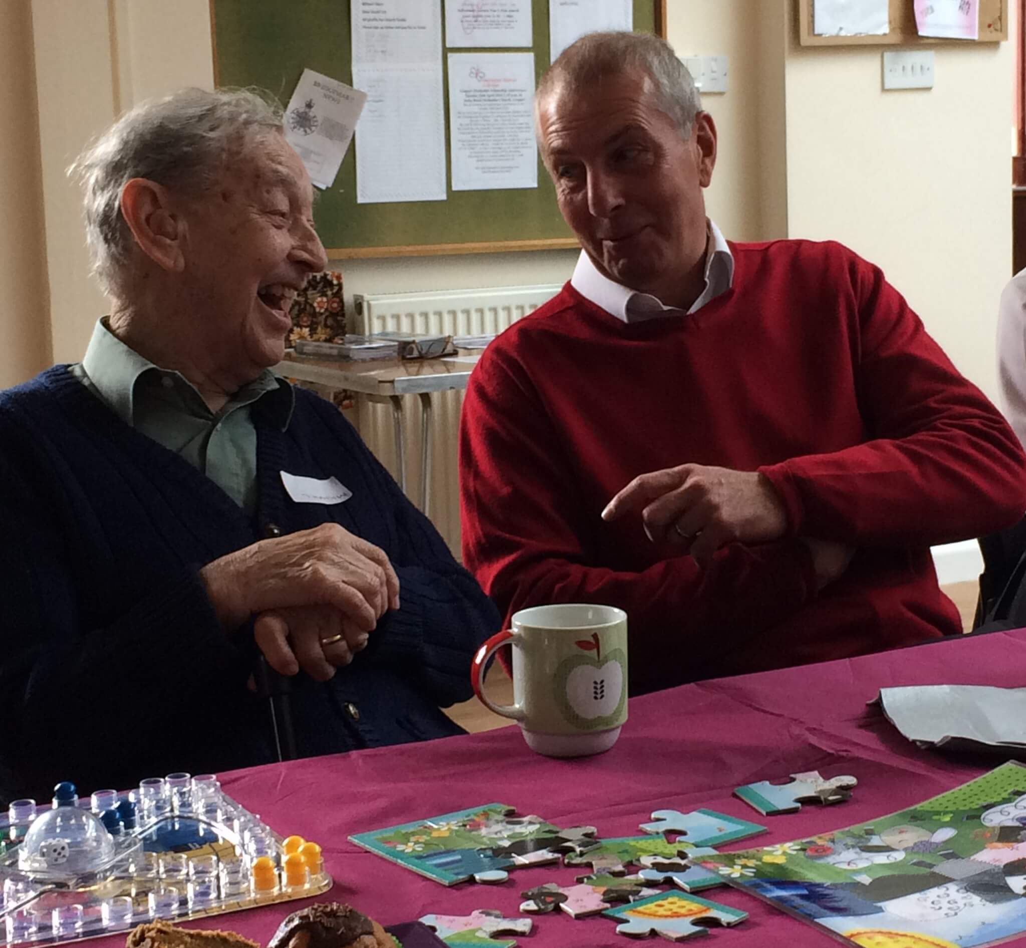 Two elderly men sit at a table with puzzles and a mug, engaged in a lively and cheerful conversation. - Home Instead