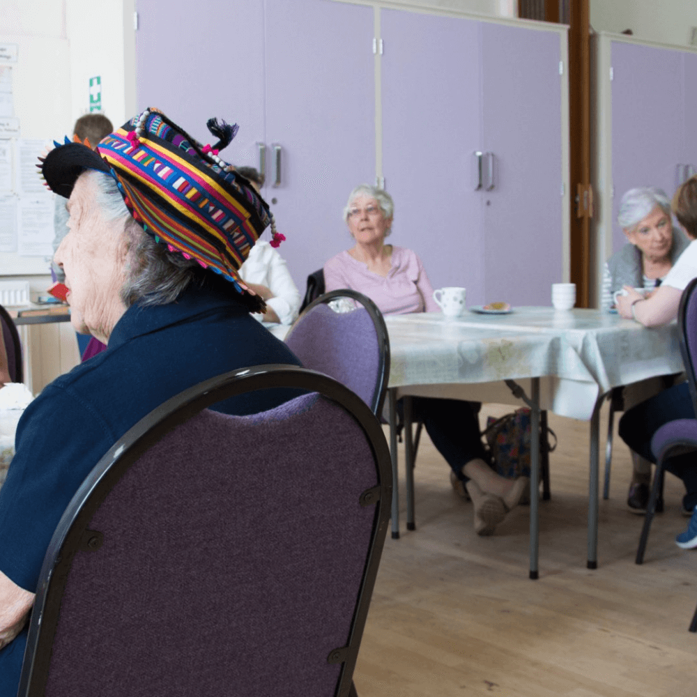 A group of elderly women, gathered in a room, engaged in conversation and activities at round tables. - Home Instead
