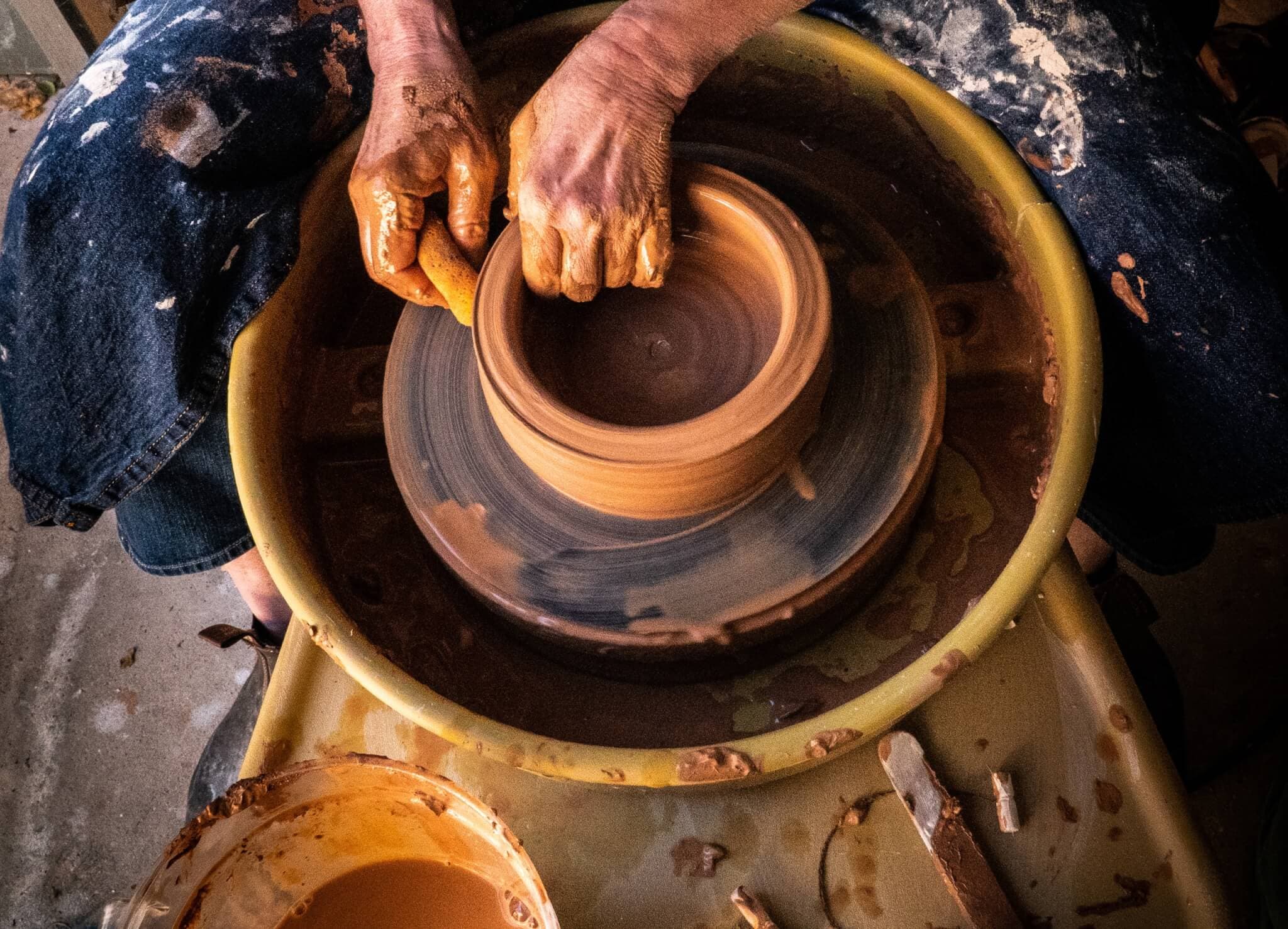 A person shapes a clay pot on a pottery wheel, with clay-covered hands and tools nearby. - Home Instead