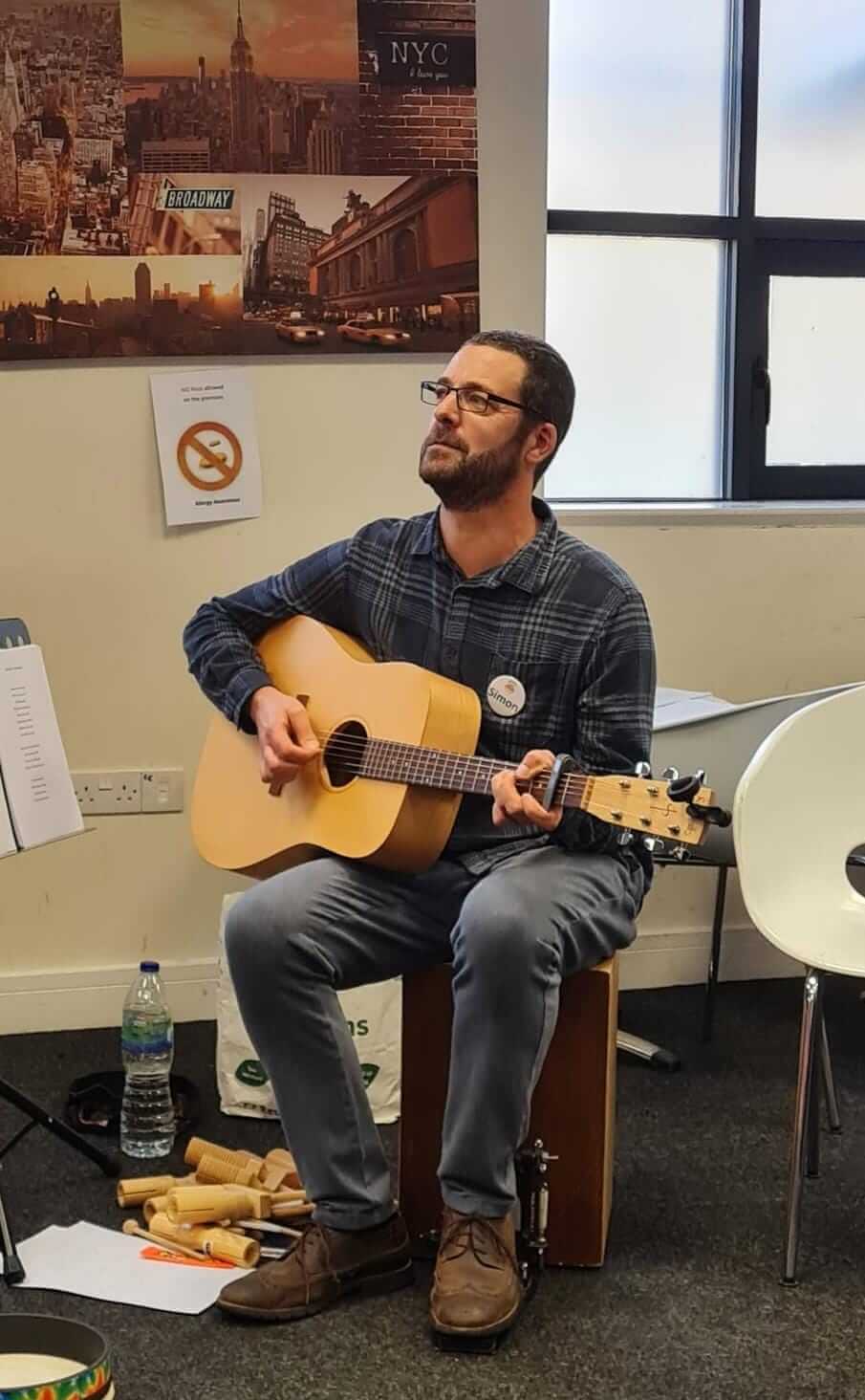 A man sits on a stool playing an acoustic guitar, with posters and a "no smoking" sign in the background. - Home Instead