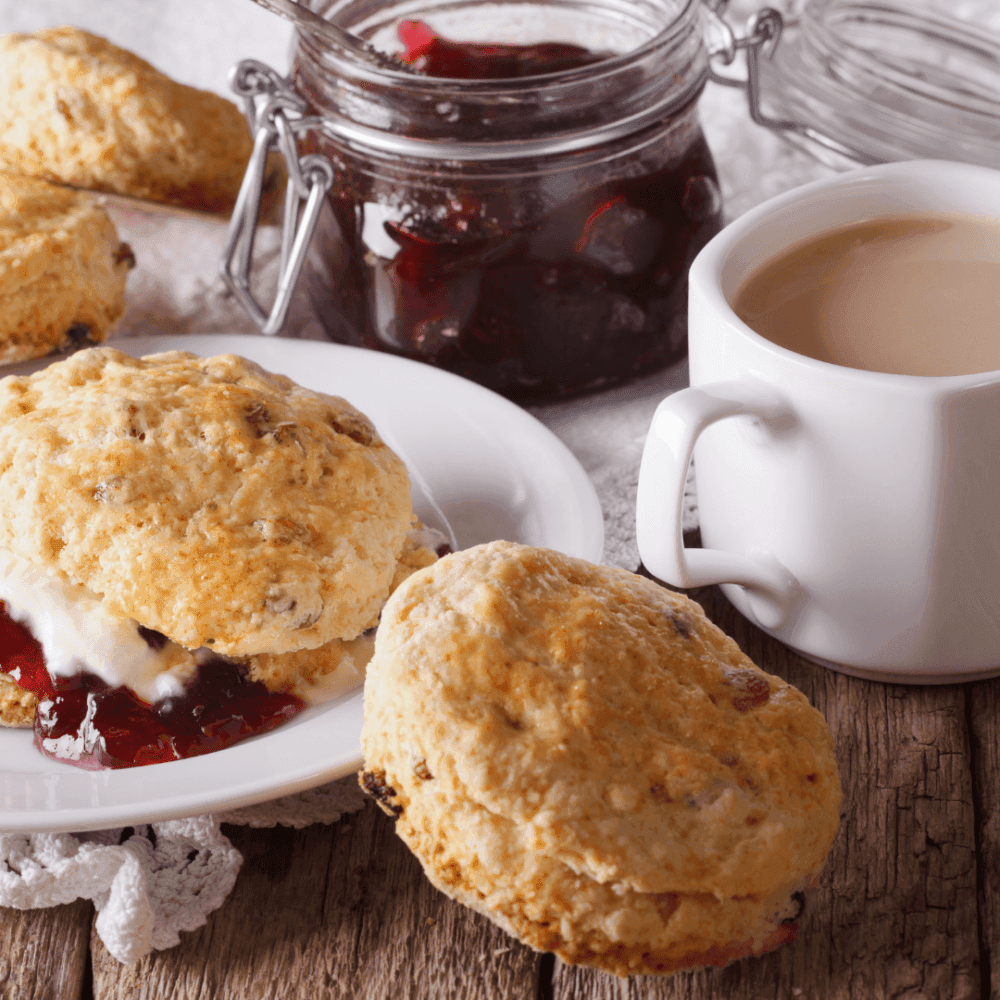 Scones with cream and jam on a wooden table, accompanied by a cup of tea and a jar of jam in the background. - Home Instead