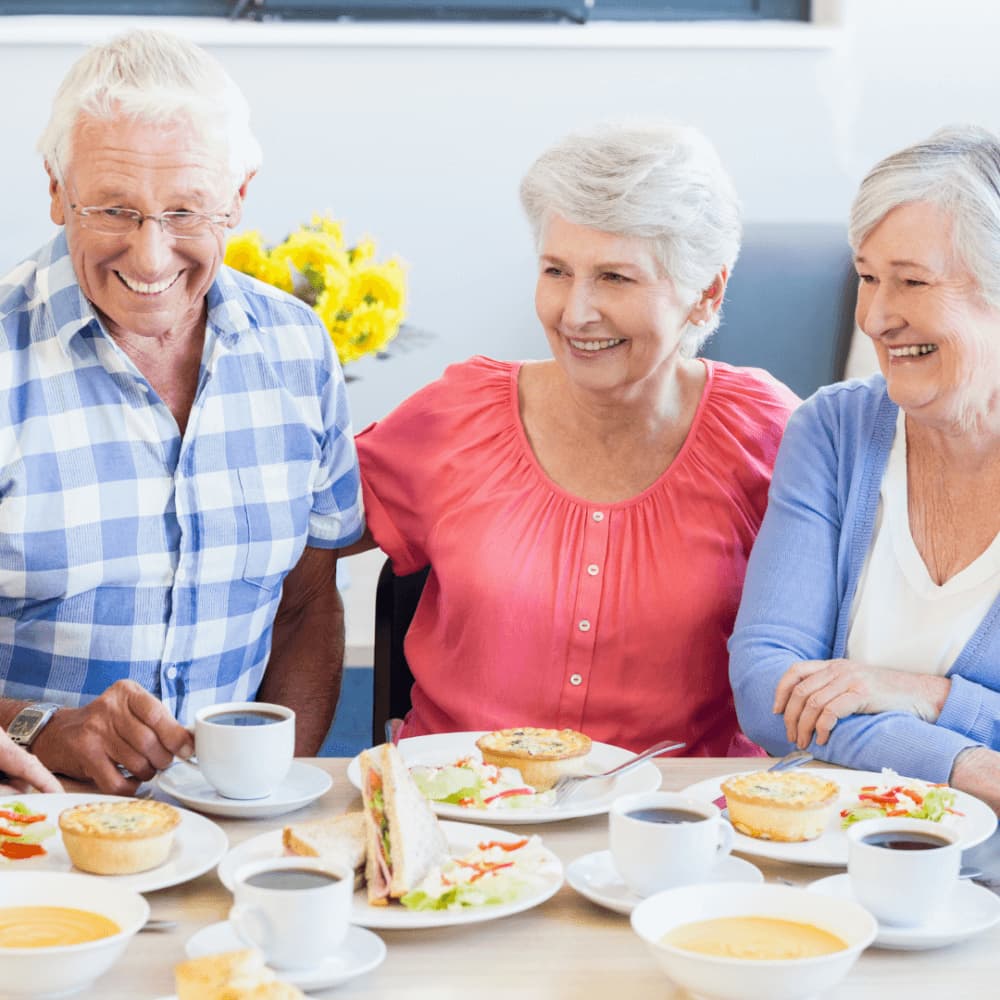 Three older adults enjoying coffee and pastries while smiling and chatting at a table. - Home Instead