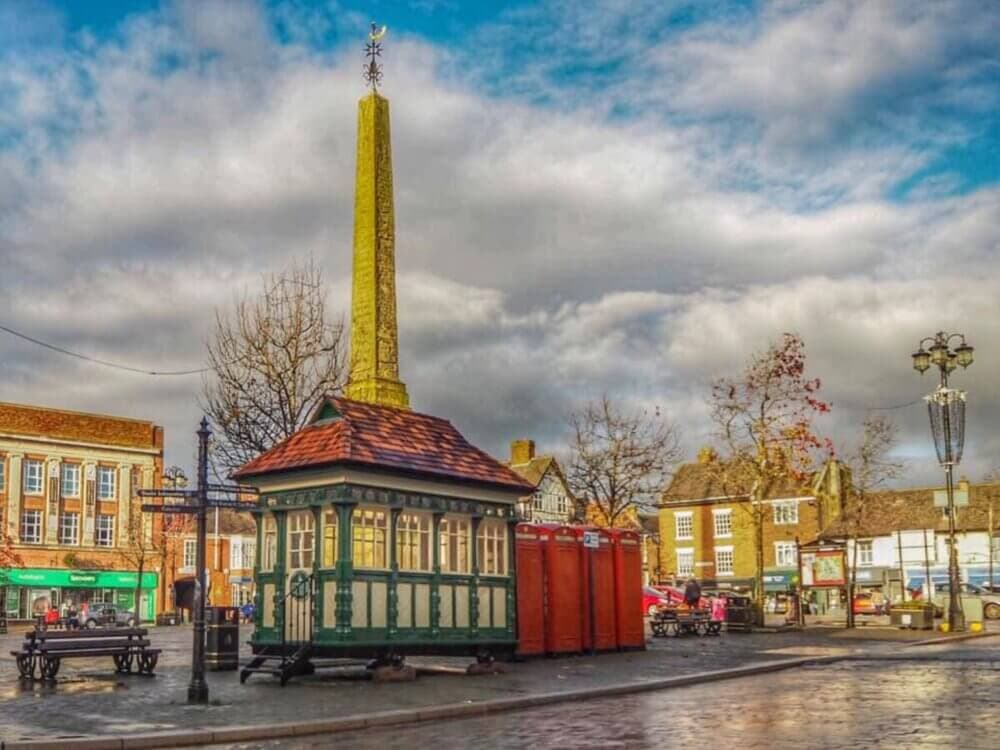 A town square with a small building, an obelisk, and red phone boxes on a cloudy day. - Home Instead