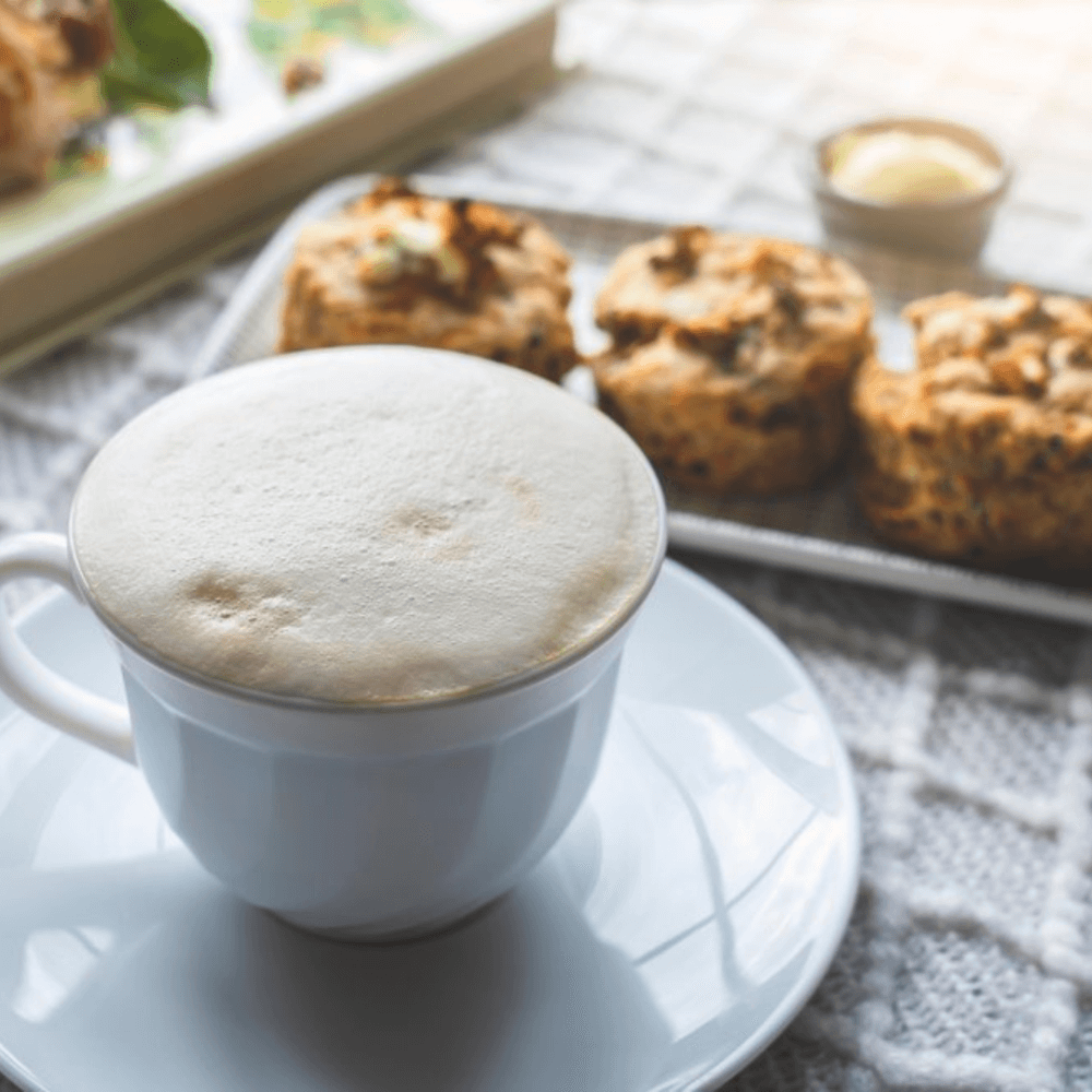 A frothy cup of coffee on a saucer in the foreground, with a tray of scones and a small dish of butter in the background. - Home Instead