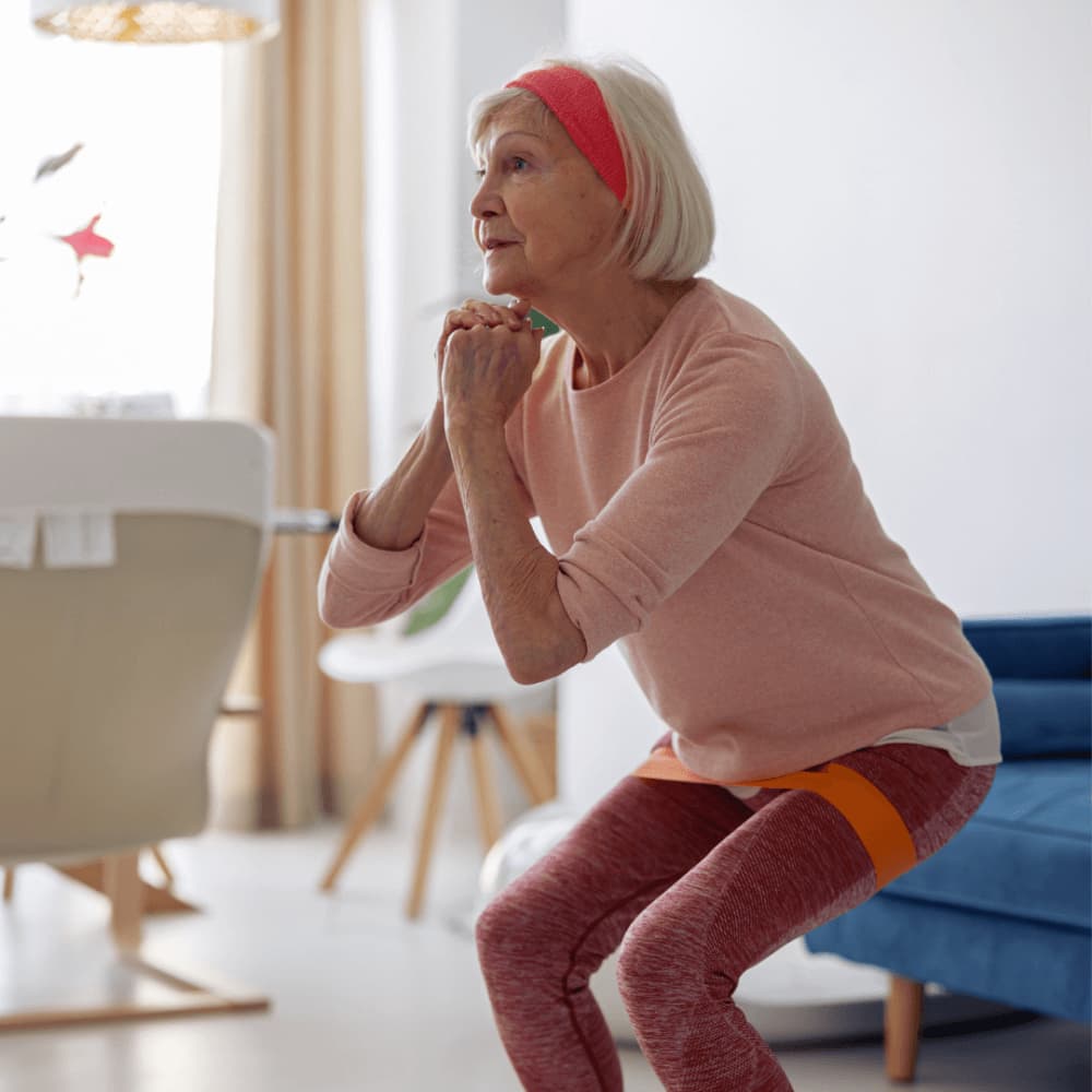 Elderly woman in athletic wear doing squats indoors with an orange resistance band around her thighs. - Home Instead