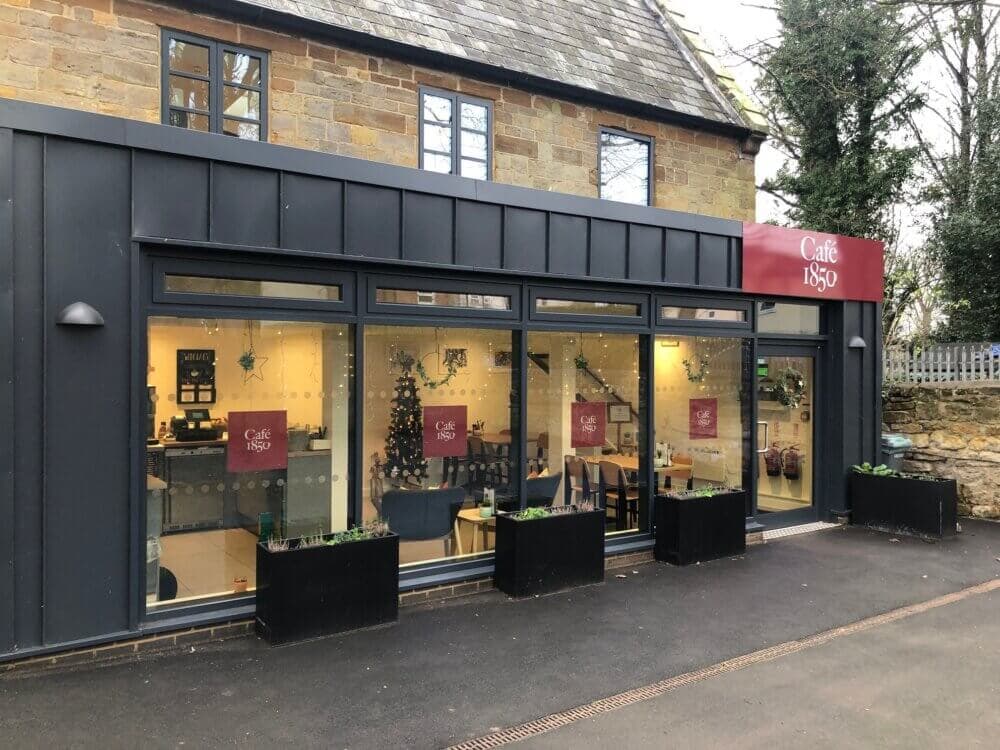 Exterior view of Café 1850 with large windows, a red sign, and festive holiday decorations inside. - Home Instead
