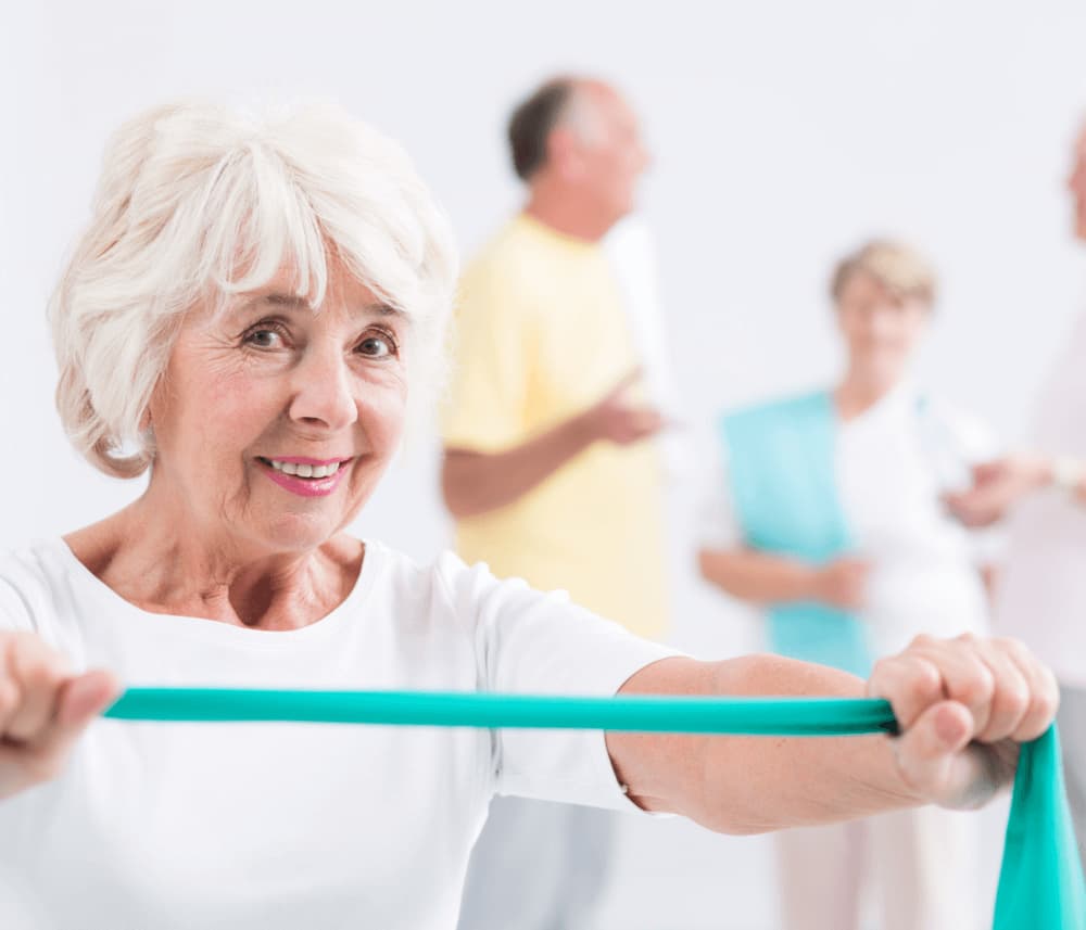Elderly woman smiling and holding an exercise band, with two men and another woman chatting in the blurred background. - Home Instead