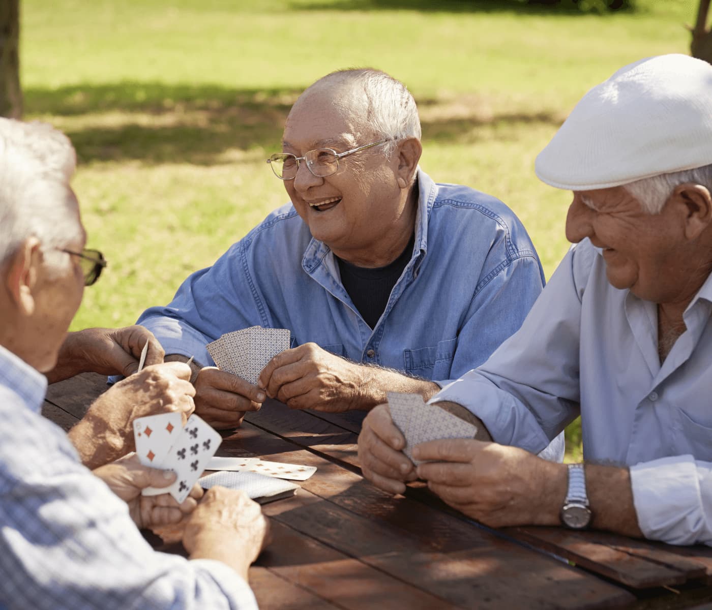 Four elderly men happily playing cards outdoors at a wooden table on a sunny day. - Home Instead