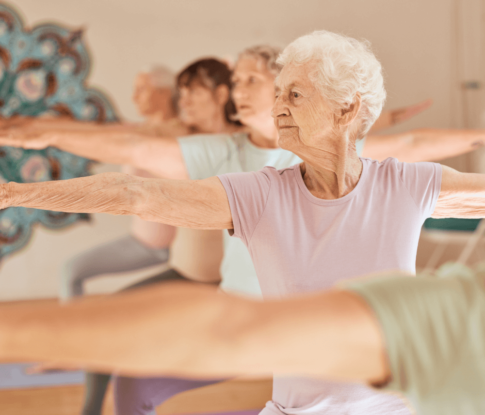 Elderly woman in a light purple shirt practicing yoga in a class, stretching her arms in a warrior pose. - Home Instead