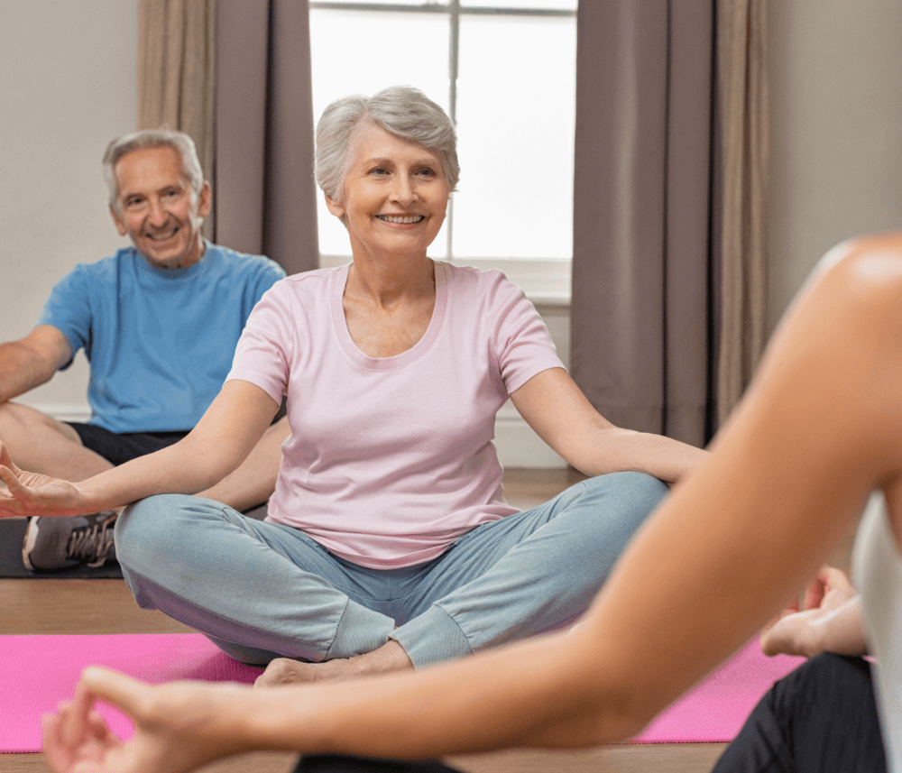 Smiling elderly couple in workout clothes sitting cross-legged on yoga mats during a group yoga session. - Home Instead