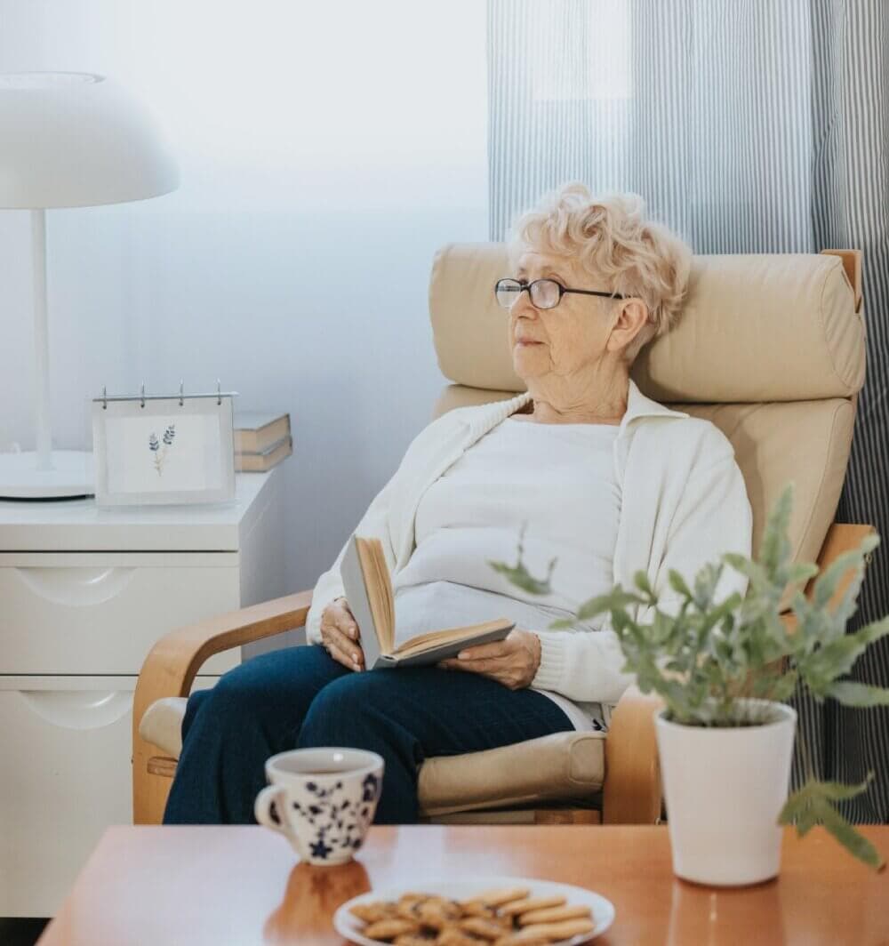 Elderly woman with glasses reading a book, sitting in an armchair with a table, cookies, and a cup beside her. - Home Instead