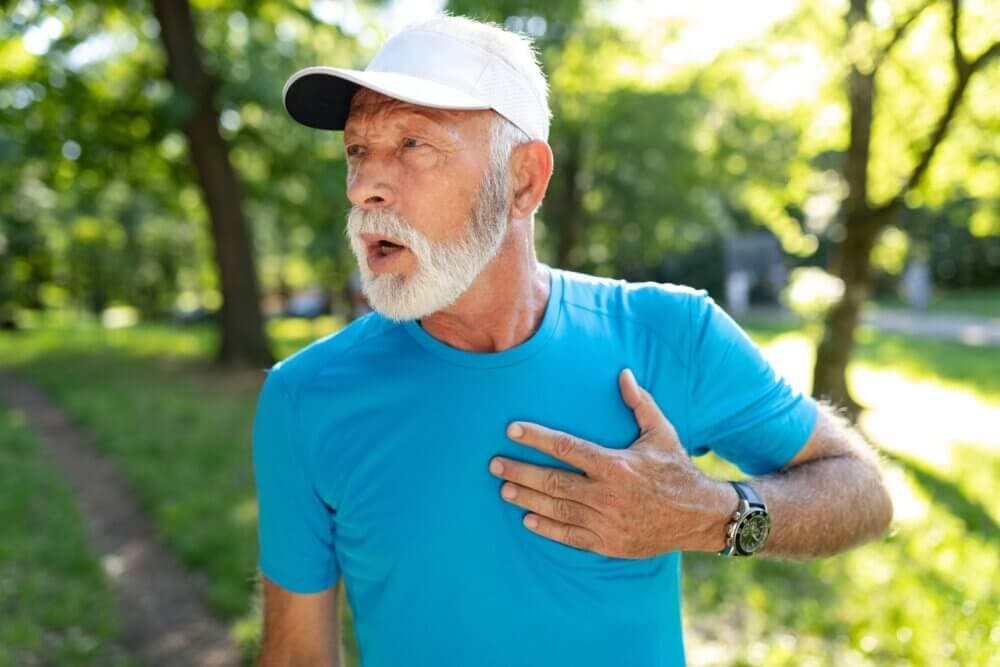 Older man in blue shirt and white cap, holding his chest and looking distressed while standing outdoors. - Home Instead
