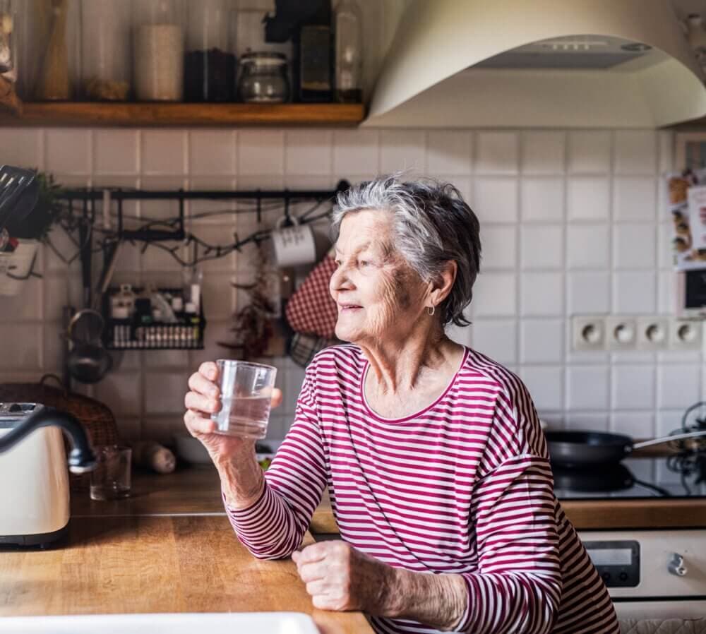 Elderly woman in a striped shirt sitting in a kitchen, smiling and holding a glass of water. - Home Instead