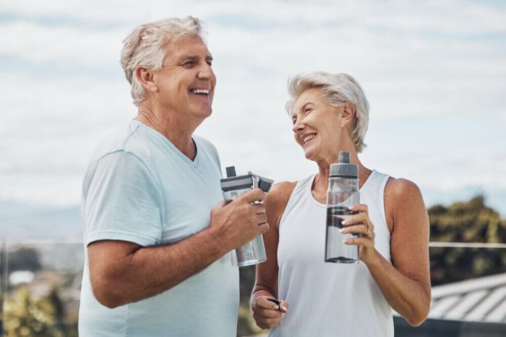 Senior couple holding water bottles and smiling at each other outdoors in athletic wear. - Home Instead