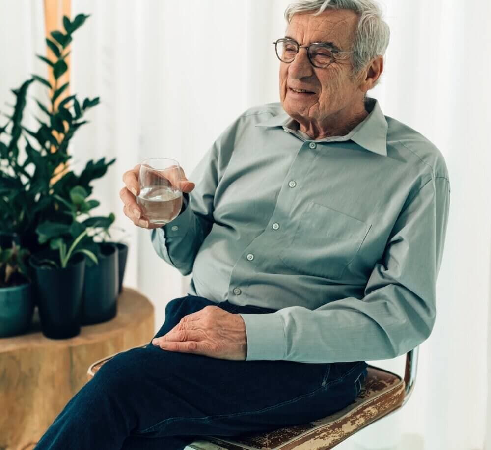 An elderly man with glasses, sitting on a chair, holding a glass of water, with plants in the background. - Home Instead