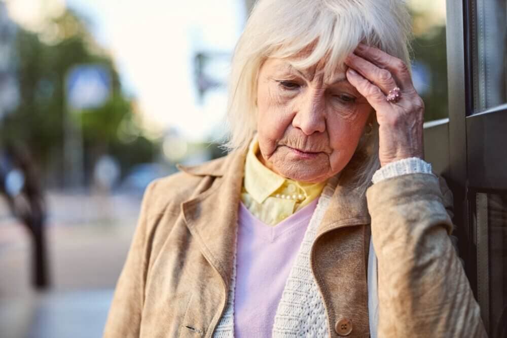 Elderly woman with white hair leaning on a wall, looking down and holding her head with a worried expression. - Home Instead