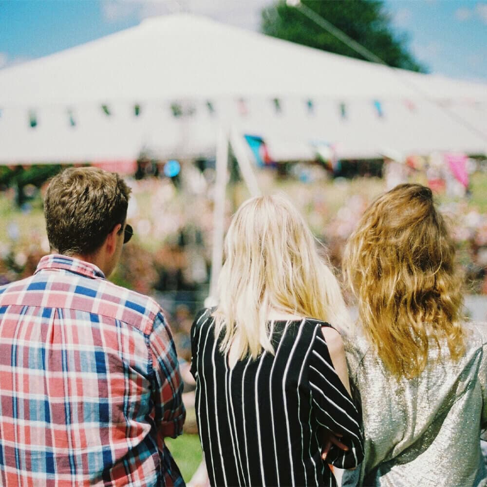 Three people with backs turned, standing outdoors at a festival with a large white tent and colorful flags. - Home Instead