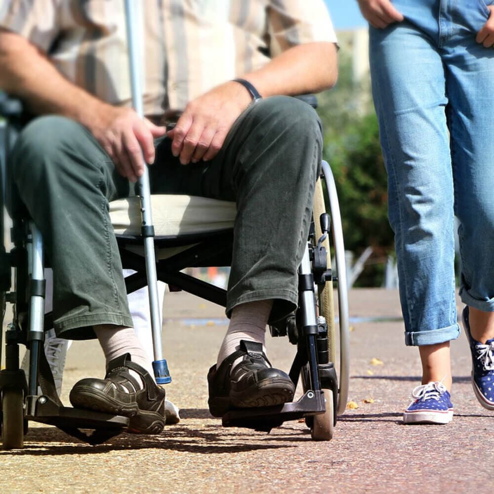 A person in a wheelchair holding a cane beside another person in jeans and sneakers, walking outdoors. - Home Instead