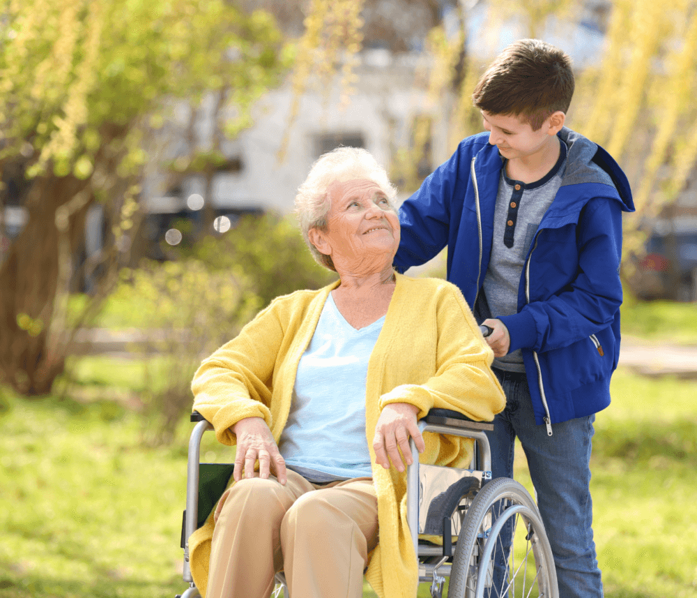 A young boy in a blue jacket pushing a smiling elderly woman in a yellow cardigan in a wheelchair outside. - Home Instead
