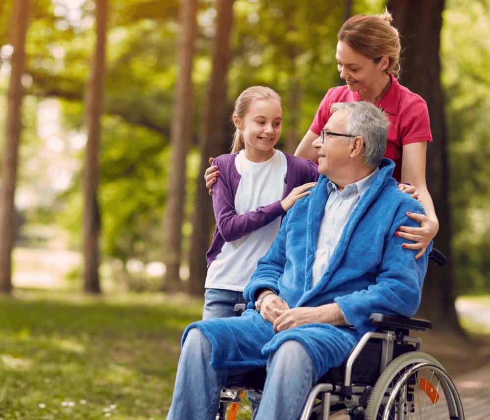 A girl and a woman smile at an elderly man in a wheelchair, sharing a moment together in a park. - Home Instead