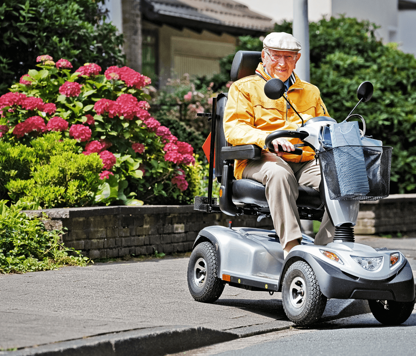 An elderly man in a yellow jacket and beige cap rides a mobility scooter on a sunny day, with blooming flowers nearby. - Home Instead