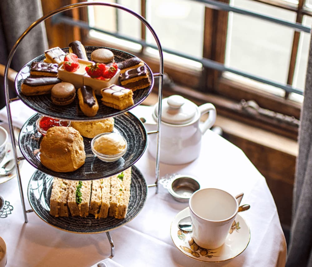 Three-tiered afternoon tea stand with assorted treats, teapot, and a cup on a white tablecloth by a window. - Home Instead
