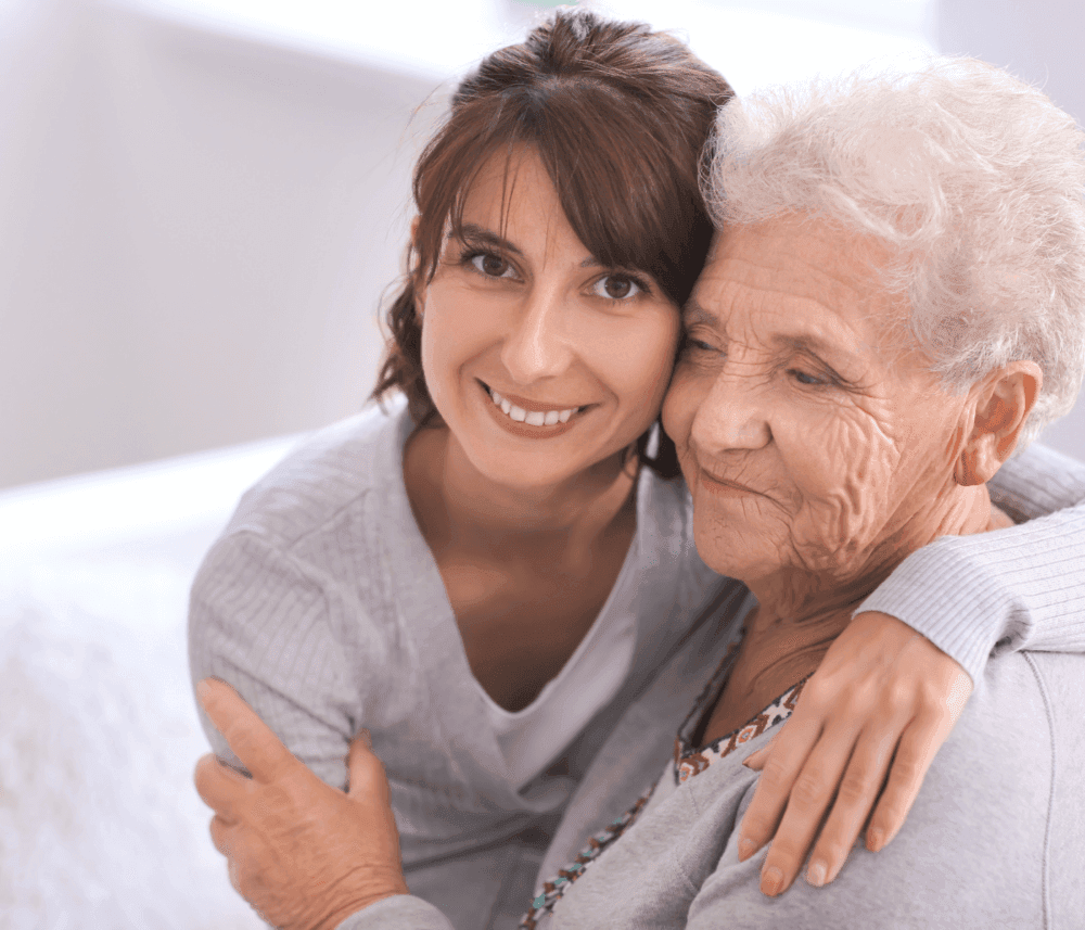 A young woman hugs an elderly woman affectionately, both smiling warmly in a light-filled room. - Home Instead