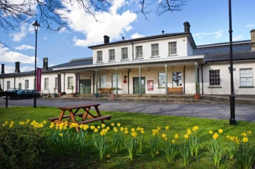 Historic white building with a porch, benches, daffodils, and a picnic table in front on a sunny, partly cloudy day. - Home Instead