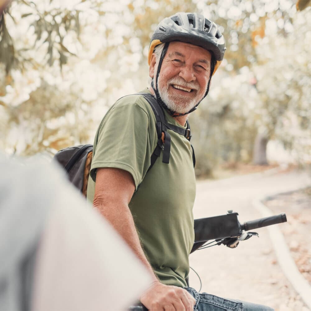 Older man smiling while sitting on a bike, wearing a helmet and green shirt, outdoors on a sunny day. - Home Instead