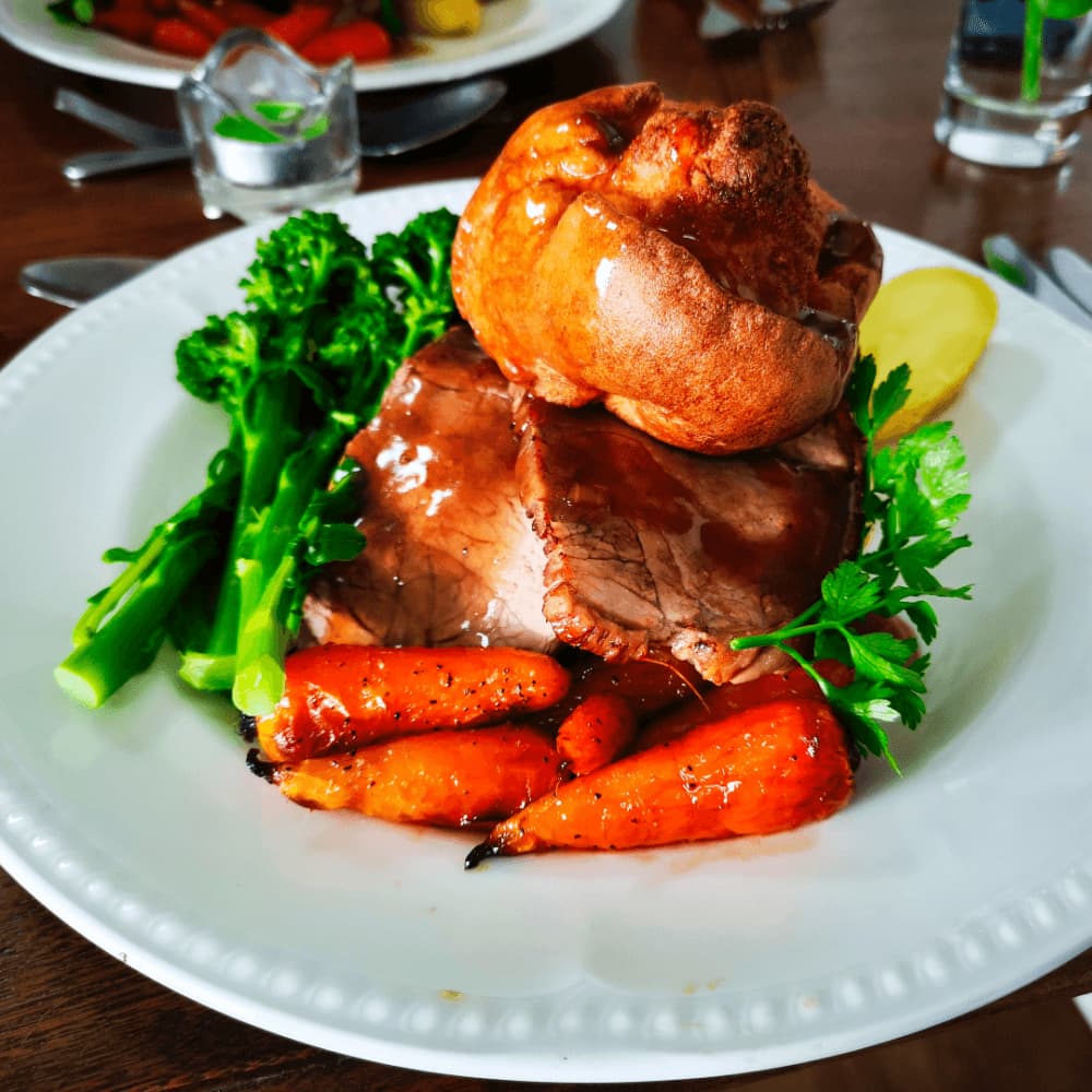 A roast dinner featuring beef, roasted carrots, broccoli, and a Yorkshire pudding on a white plate with a glass of water. - Home Instead