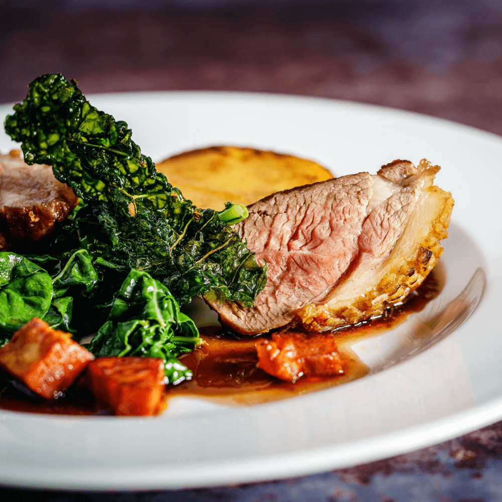 A plate of seared meat with crispy skin, greens, diced vegetables, and a round potato piece drizzled with sauce. - Home Instead