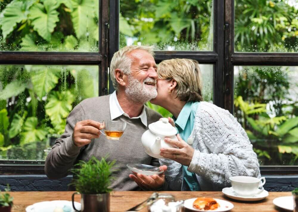 Elderly couple enjoying tea, woman kissing man's cheek, seated in front of window with lush greenery outside. - Home Instead
