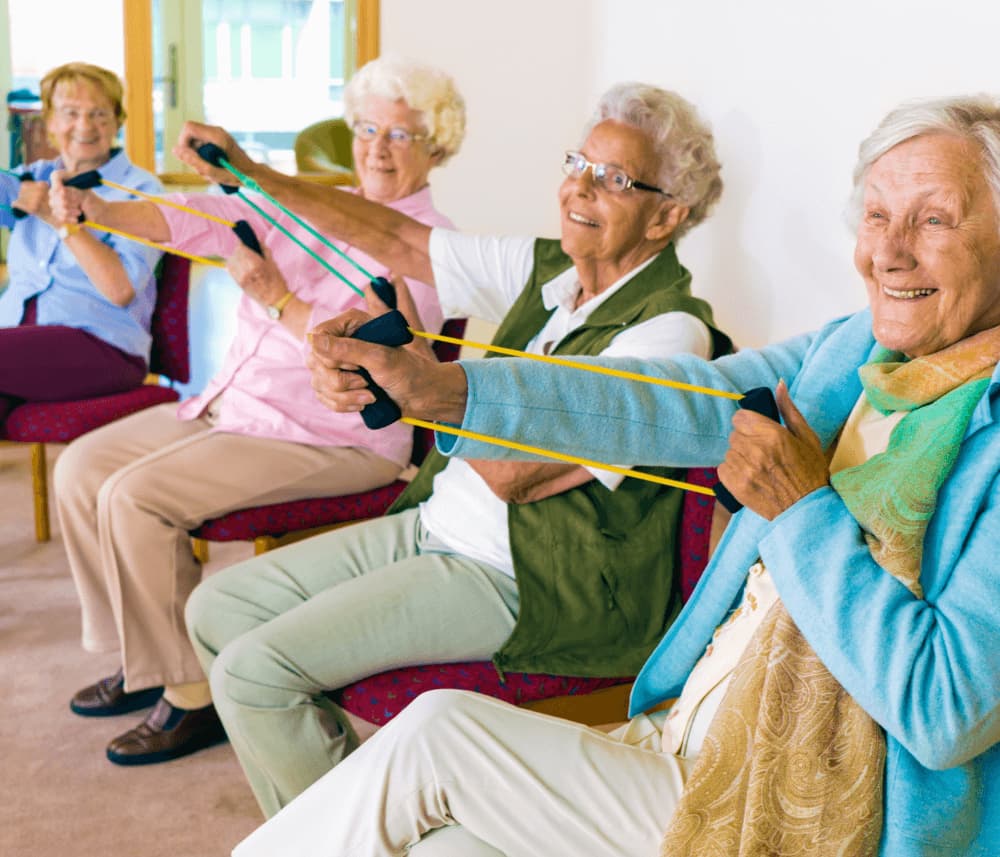 Four elderly women seated in chairs, smiling while using resistance bands for exercise in a bright room. - Home Instead