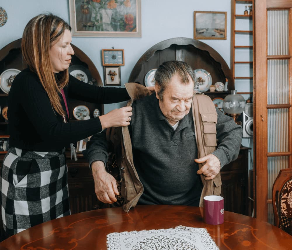 A woman helps an elderly man put on a jacket while he stands beside a table with a pink mug and lace doily. - Home Instead