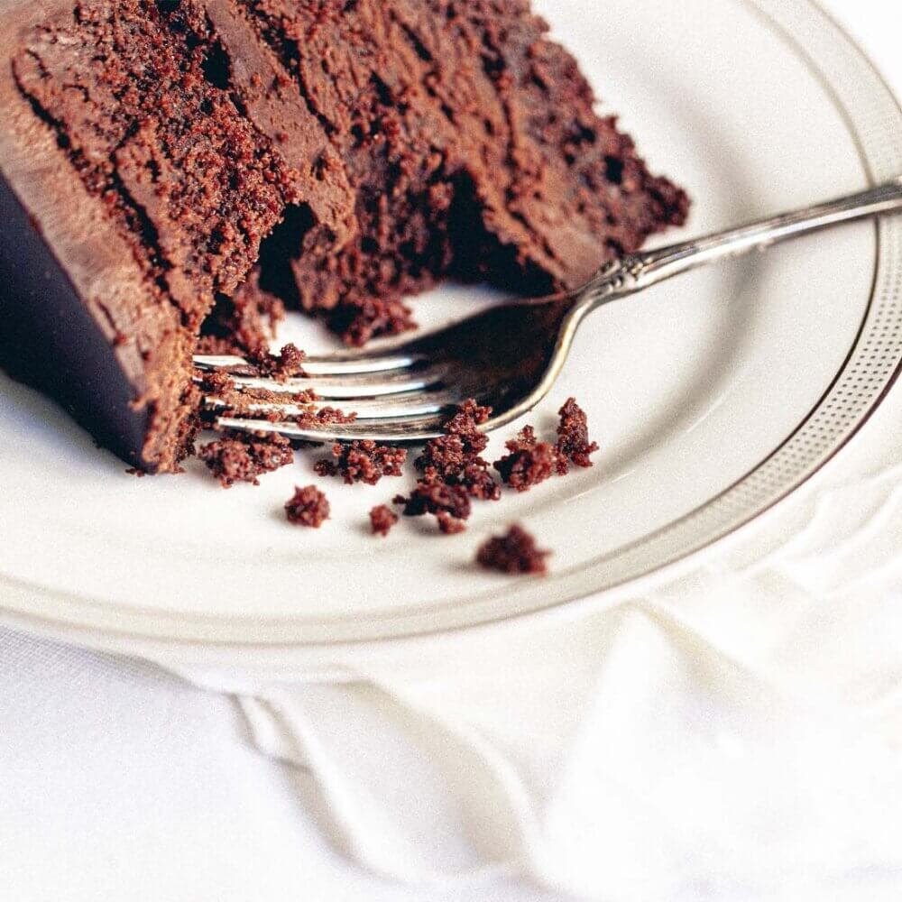 A slice of rich chocolate cake on a white plate, with a fork partially inserted into the cake and some crumbs around it. - Home Instead