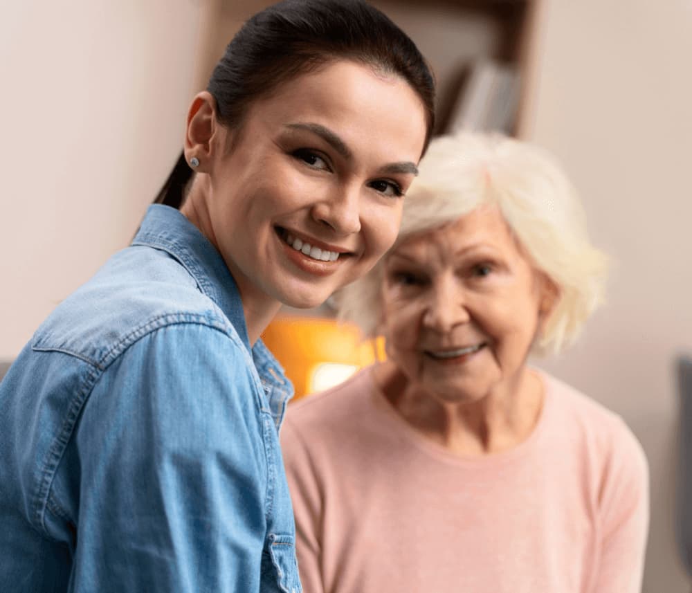 Young woman smiling at camera next to older woman in pink sweater, both looking happy indoors. - Home Instead