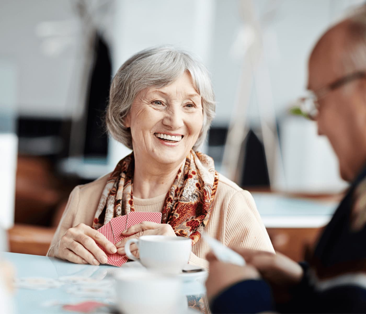 Smiling elderly woman holding playing cards, sitting at a table with a cup of coffee, conversing with a man. - Home Instead