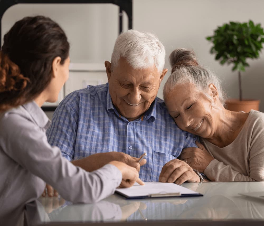 An elderly couple smiles and leans together as they sit with a younger woman reviewing documents at a table. - Home Instead