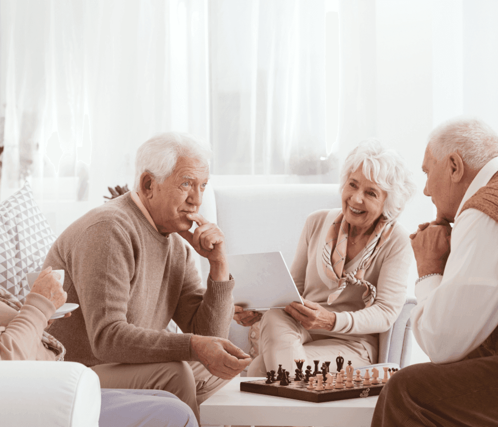 Elderly friends gathered in a cozy living room, smiling and chatting, with a chessboard in front of them. - Home Instead