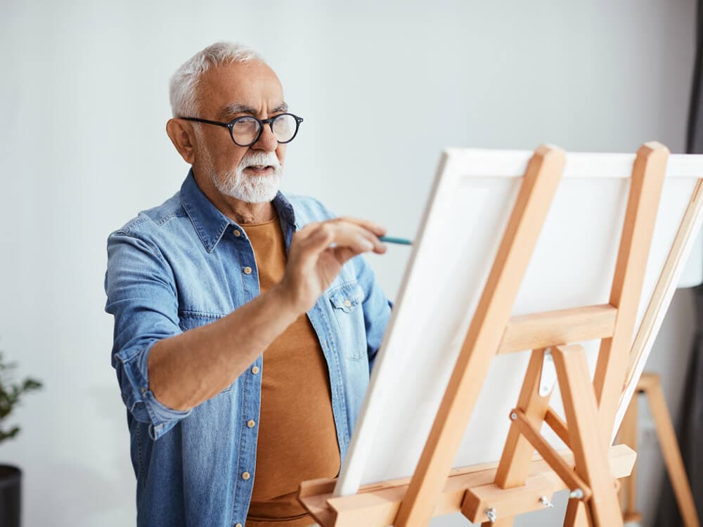 Elderly man with glasses and a denim shirt painting on a canvas at an easel in a bright room. - Home Instead